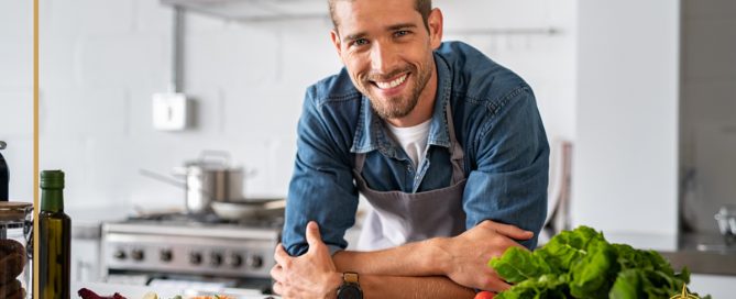 Smiling man in apron leaning on kitchen counter with fresh vegetables, stainless steel stove and modern range hood in the background.
