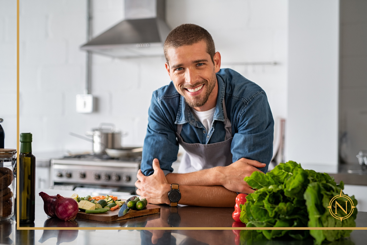 Smiling man in apron leaning on kitchen counter with fresh vegetables, stainless steel stove and modern range hood in the background.
